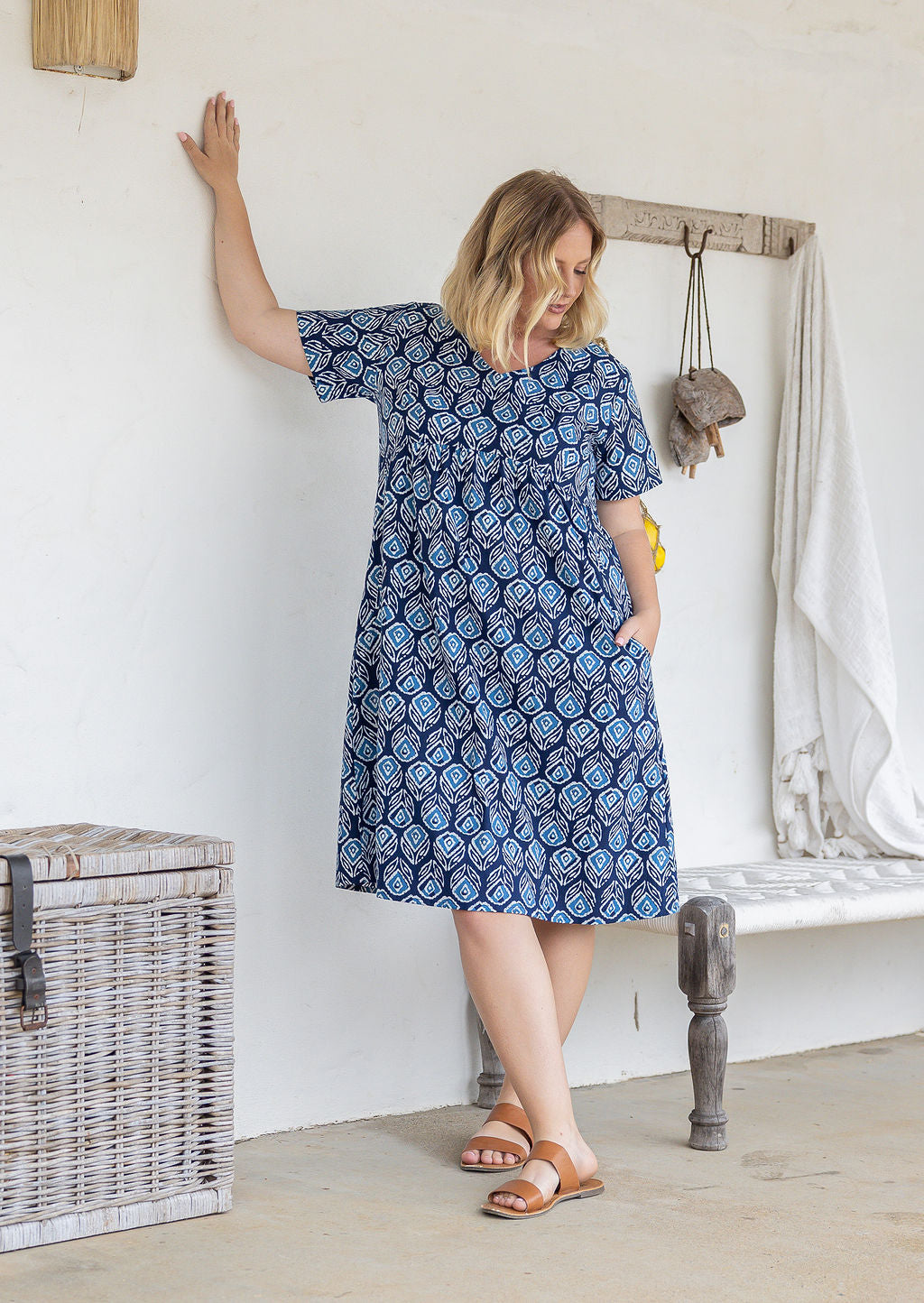 Model wearing a cotton knee length dress with empire waist, short sleeves and hand in pocket paired with brown sandals.