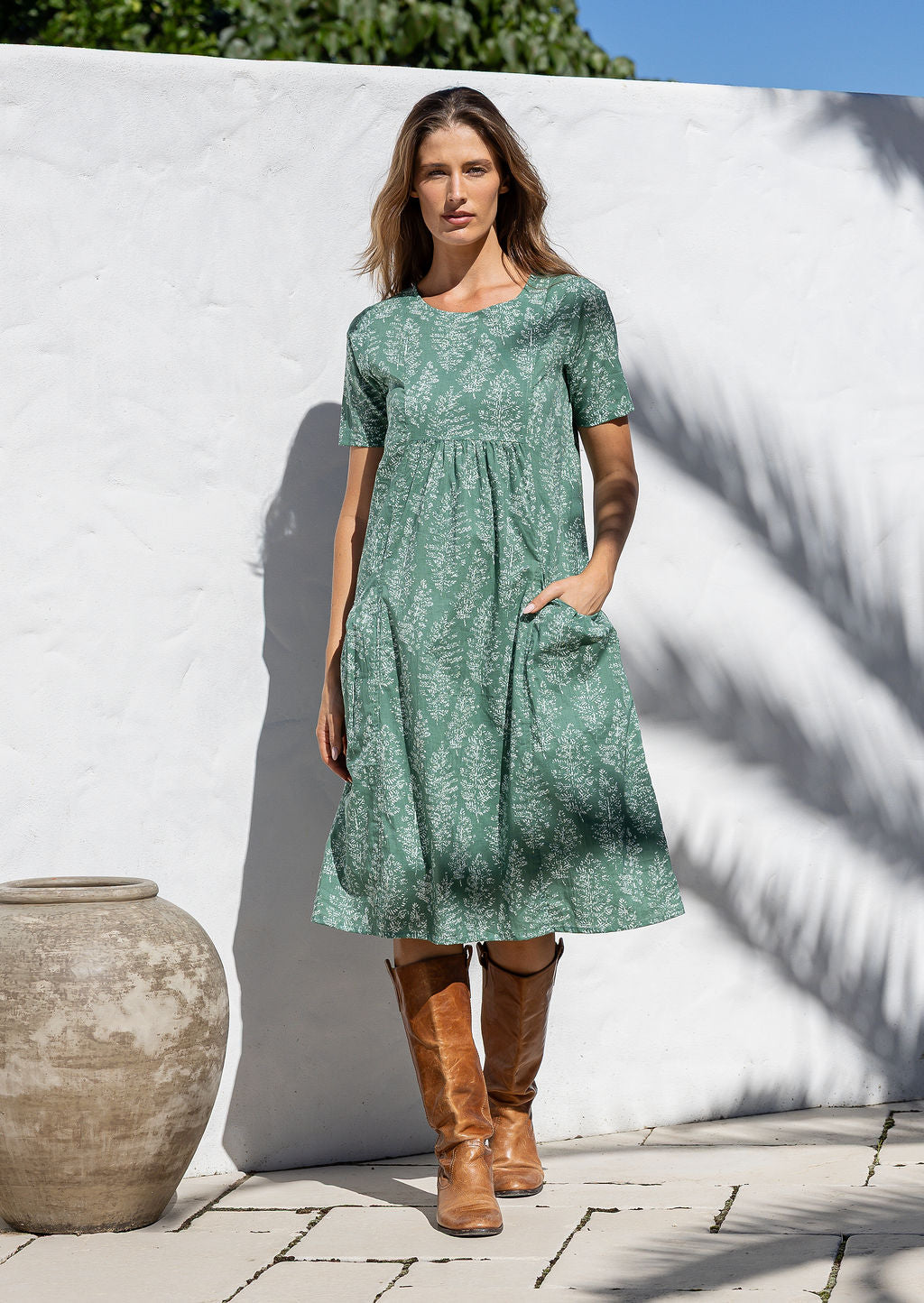 Woman wearing a light sage green cotton dress with a white wall and pot in the background
