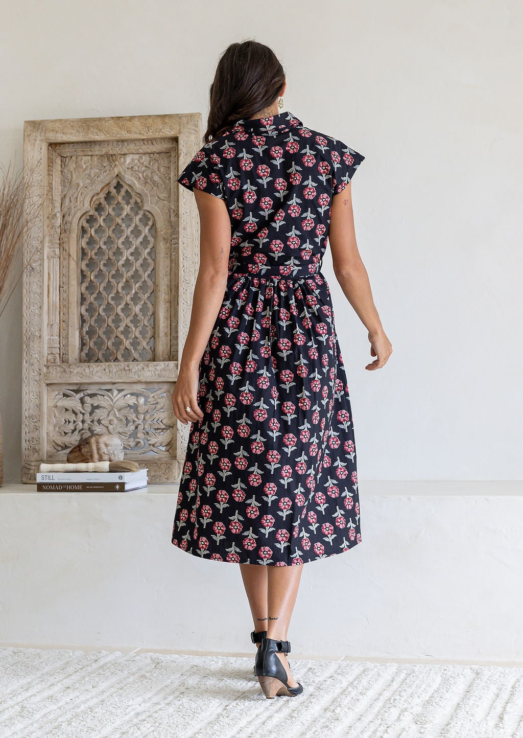 Woman stands backwards wearing a 100% cotton Black floral midi shirtdress with cap sleeves and tie waist.