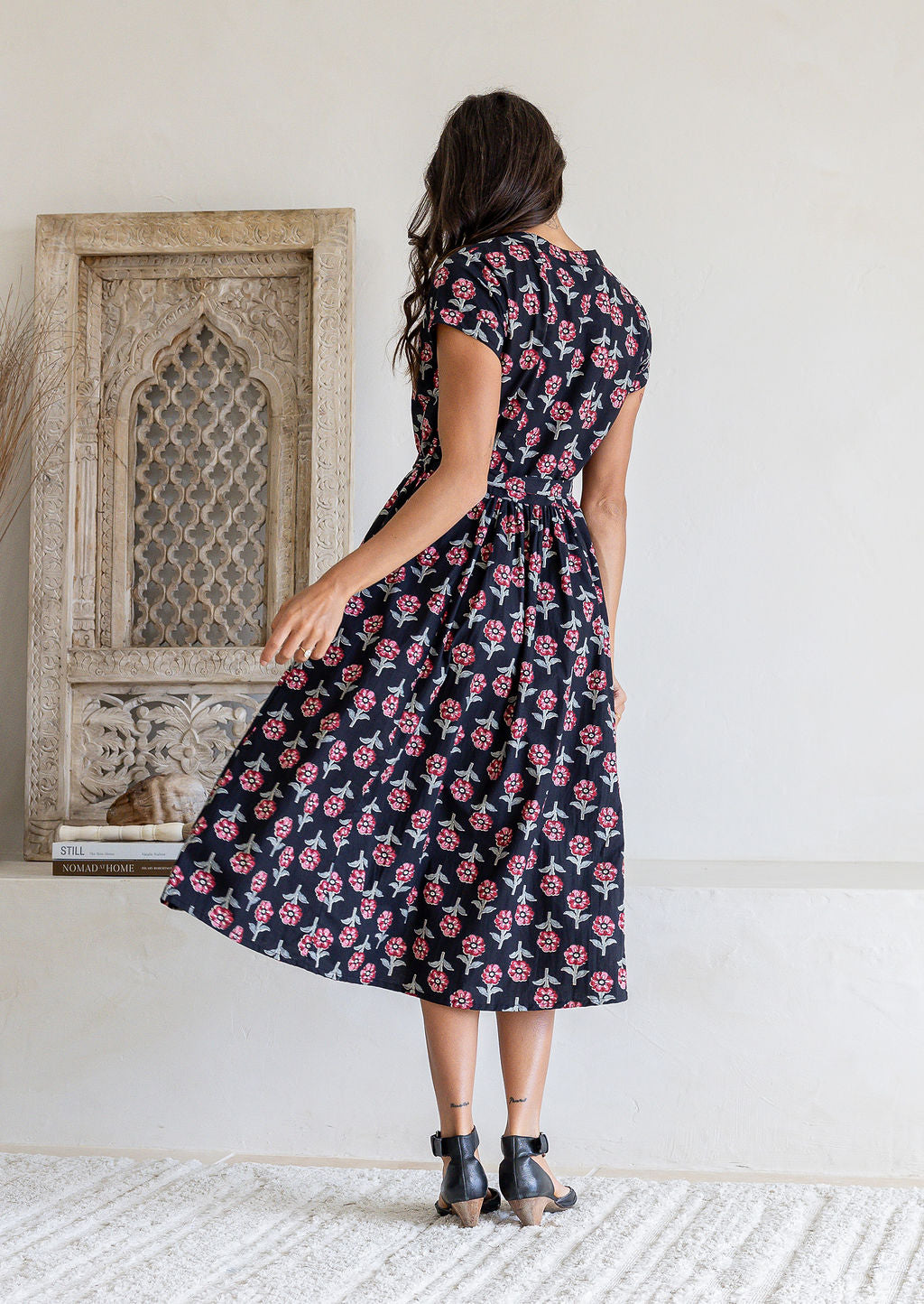 Woman stands backwards wearing a 100% cotton black based floral print midi dress with cap sleeves paired with black heels.