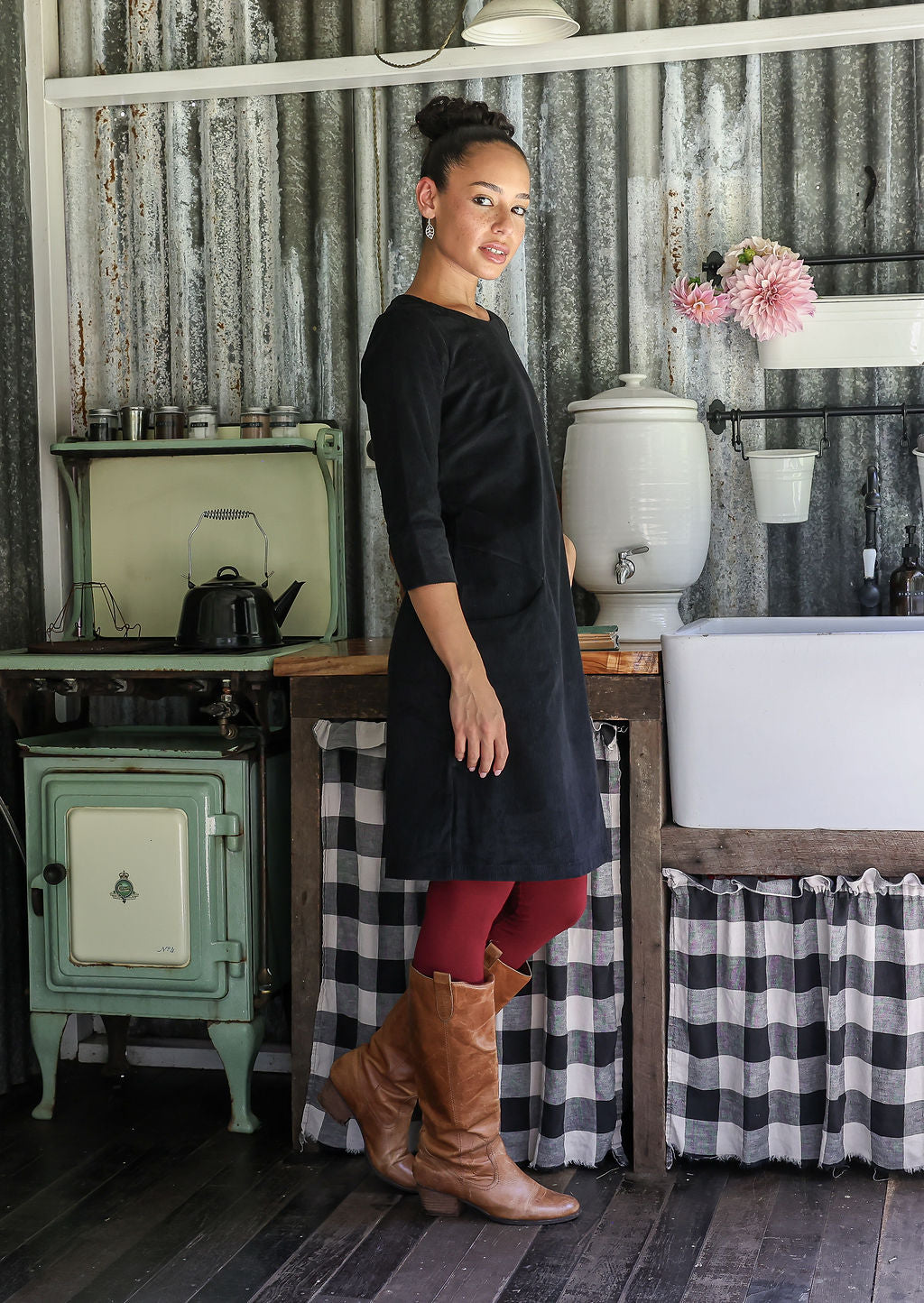 Woman standing in a rustic kitchen in a cotton corduroy black dress with 3/4 sleeves and pockets, paired with maroon leggings and tan boots