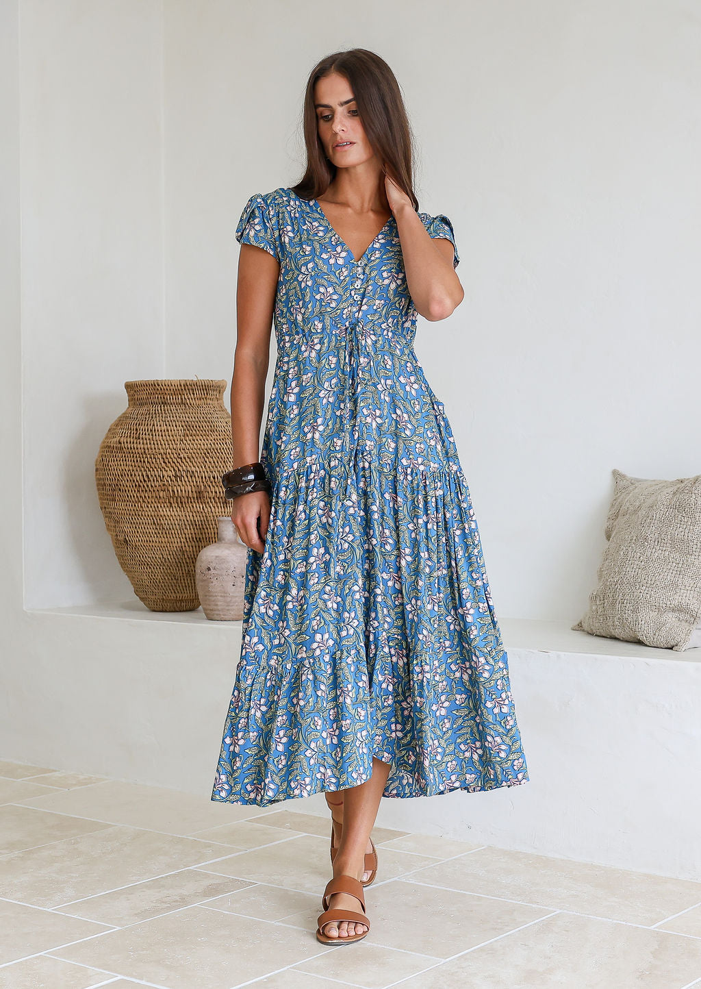 Woman poses in blue based block style floral print rayon maxi dress with V-neckline, buttoned bodice, waist-tie and pockets