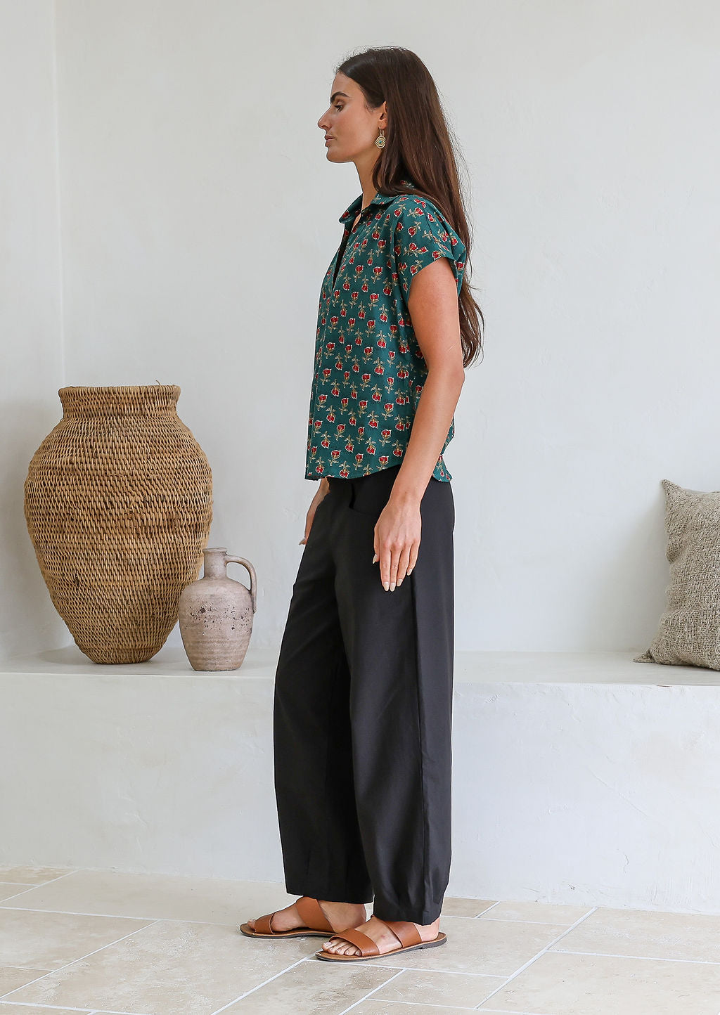 Woman stands sideways wearing a green based floral cotton top with short folded cuff sleeves, collar and V neckline