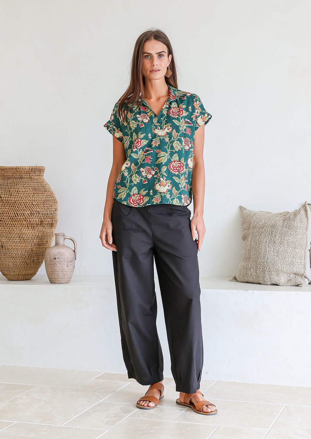 Woman wearing a green based floral print 100% cotton top and black pants in a minimal indoor setting.