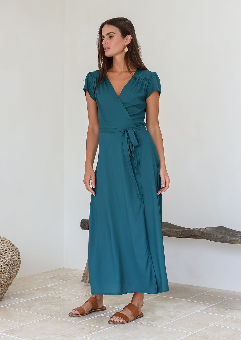 Woman wearing a teal rayon wrap maxi dress standing in a minimal indoor setting.