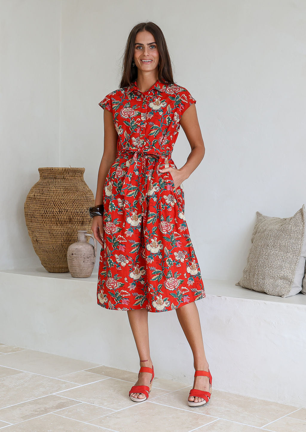 Woman wearing a midi length red based floral cotton dress with buttoned bodice, standing against a white wall and decorative vases
