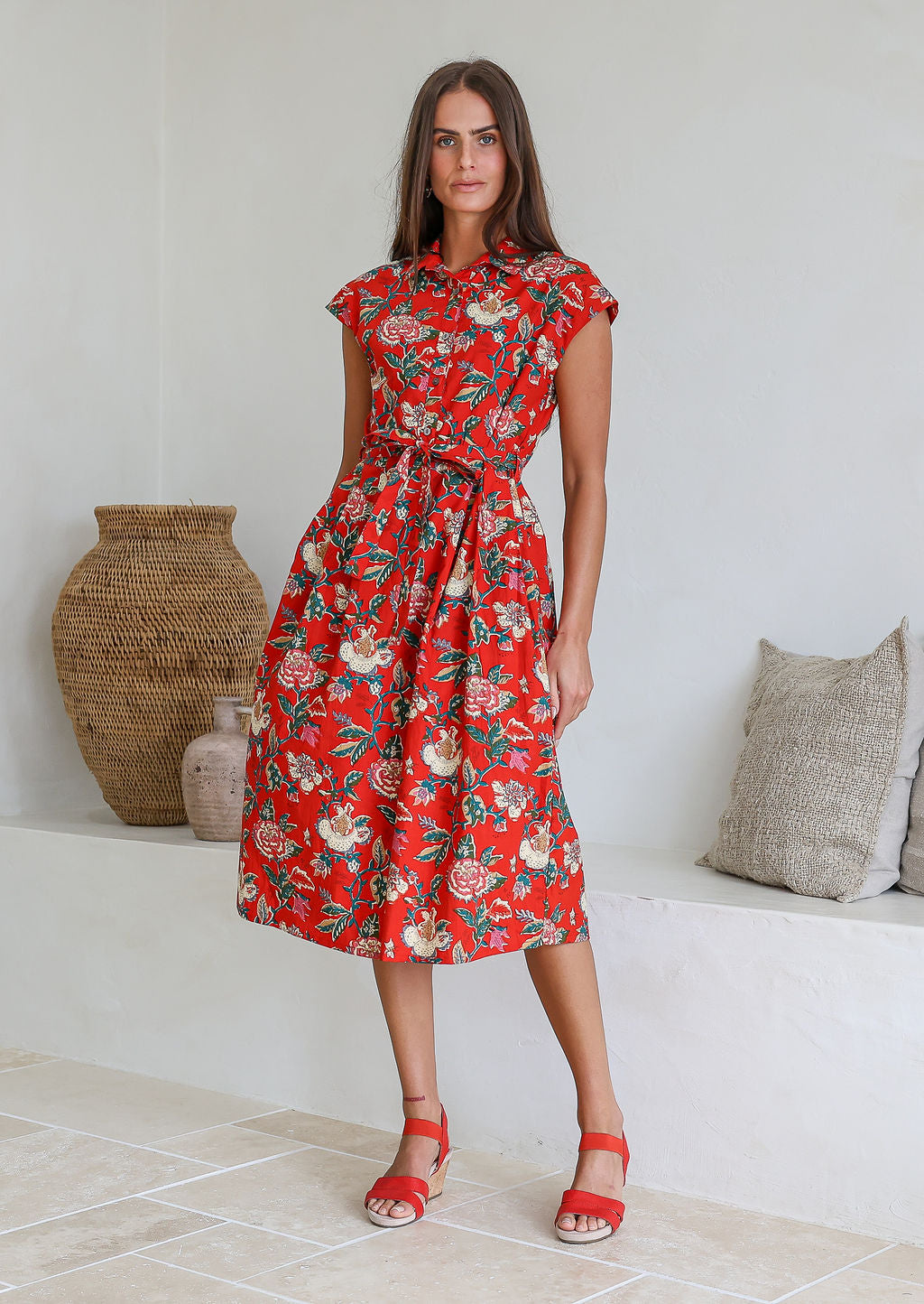 Model stands with wearing a midi length floral printed shirt style cotton dress with cap sleeves, buttoned bodice and a removable waist tie