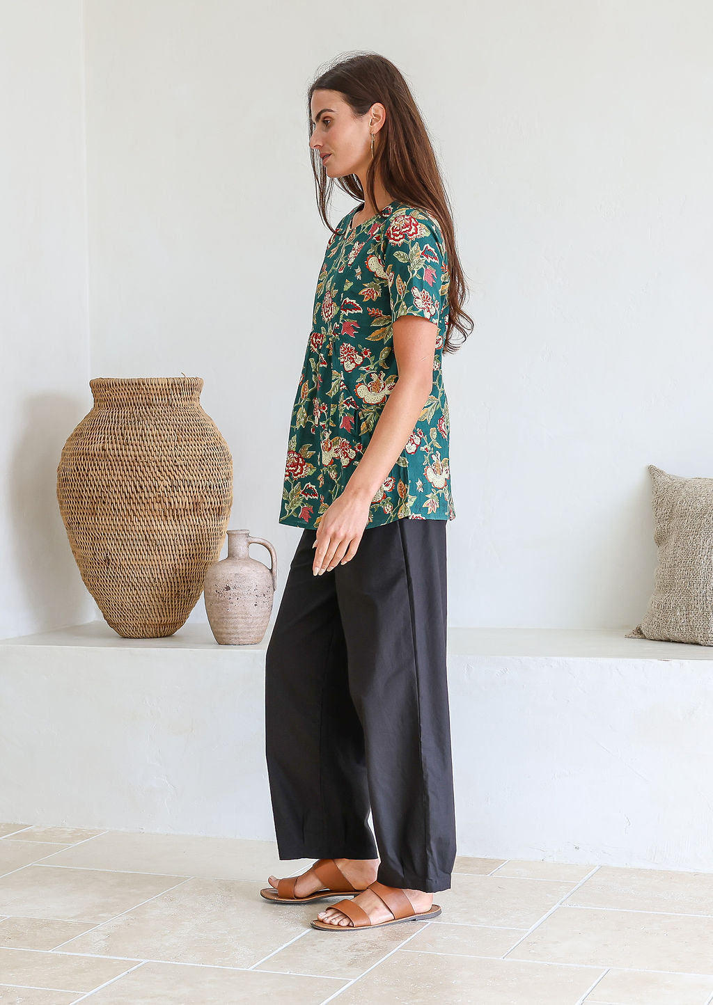 Woman stands sideways wearing a relaxed fit bottle green based floral print top against a white wall.