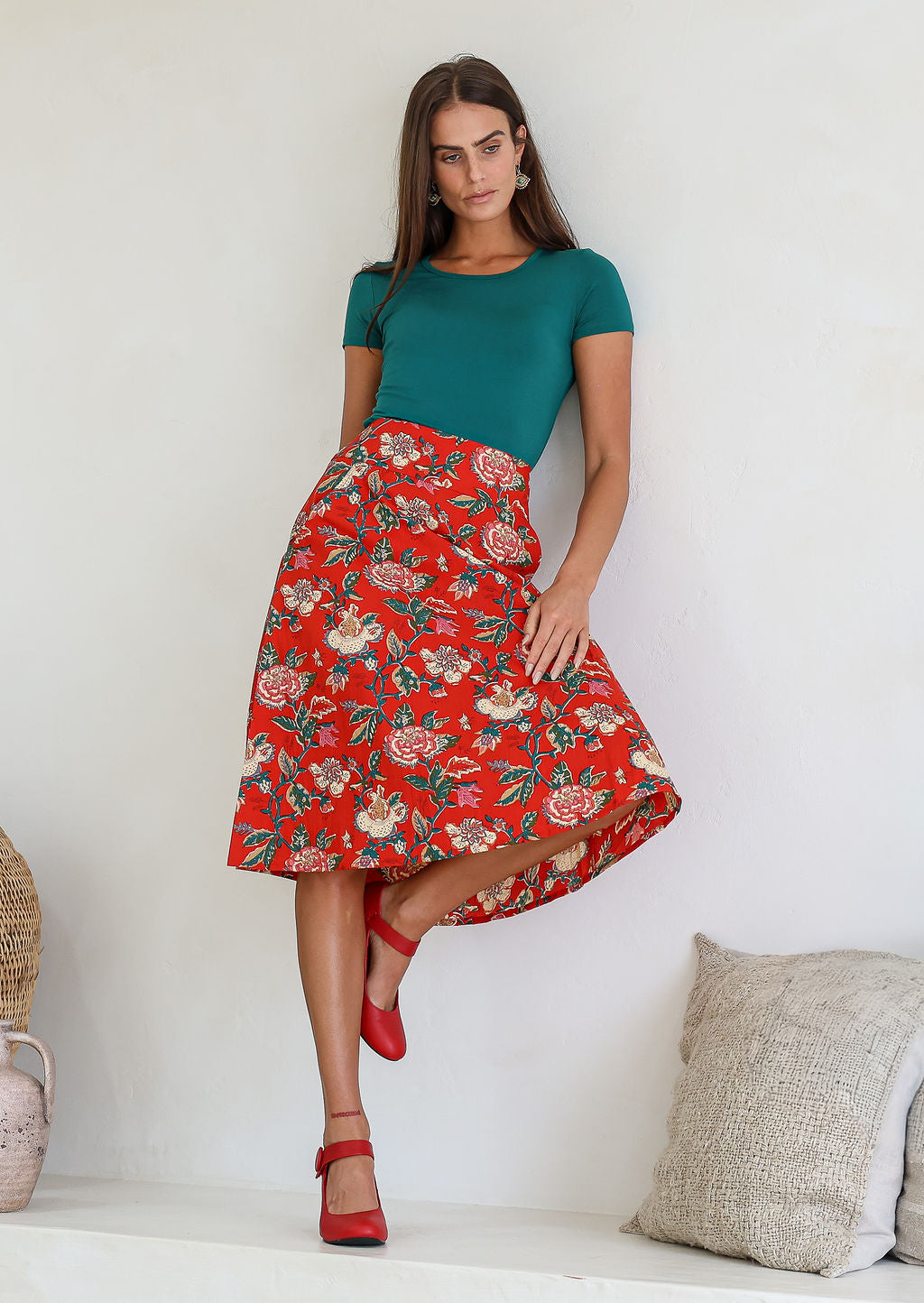woman leans on wall wearing a knee length floral cotton skirt with a-line shape, pockets and side zipper.