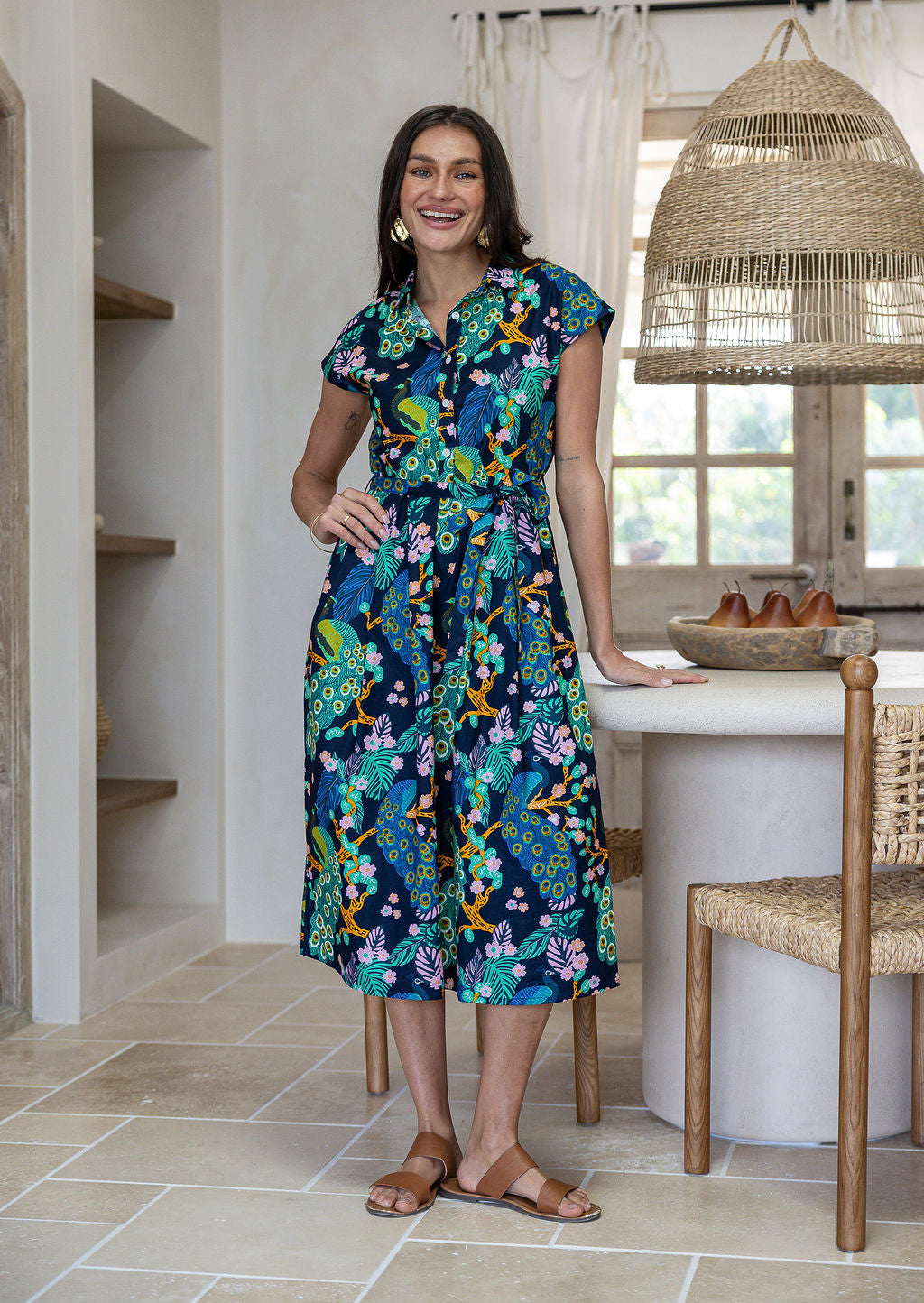 A woman wearing a cap-sleeved, collared cotton midi dress with a multicolour peacock and floral print on a dark navy base