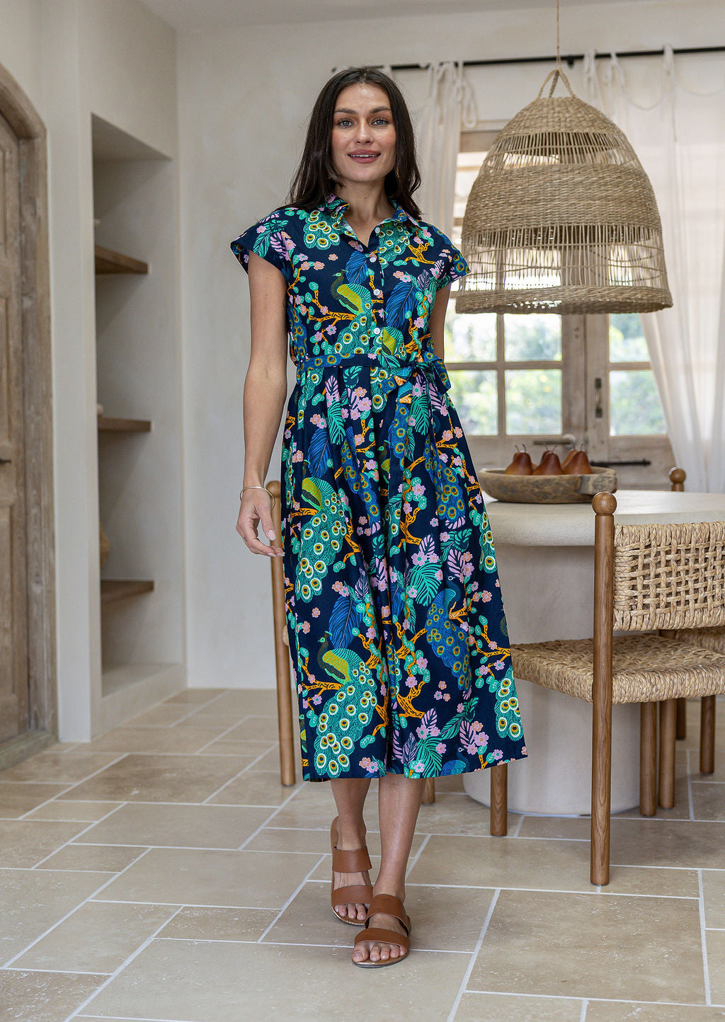 A woman modelling a cap-sleeve cotton midi dress with collar and buttoned bodice, in a vibrant multicolour floral and peacock print