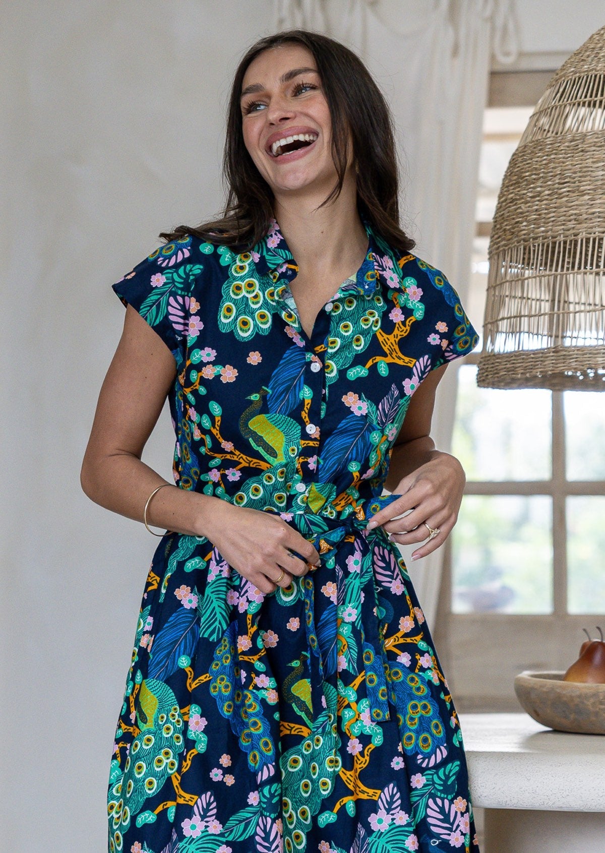 Close up of woman wearing a navy blue based peacock print cotton dress with collar, buttoned bodice and waist tie