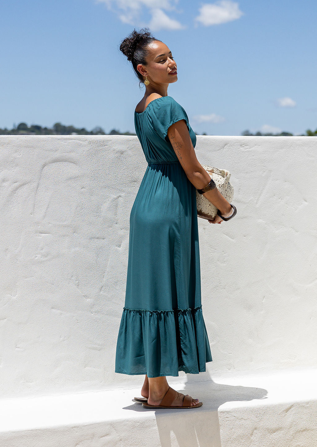 Side view of Woman in a teal maxi, perfect Christmas dress holding a ceramic pot against a white wall with blue sky.