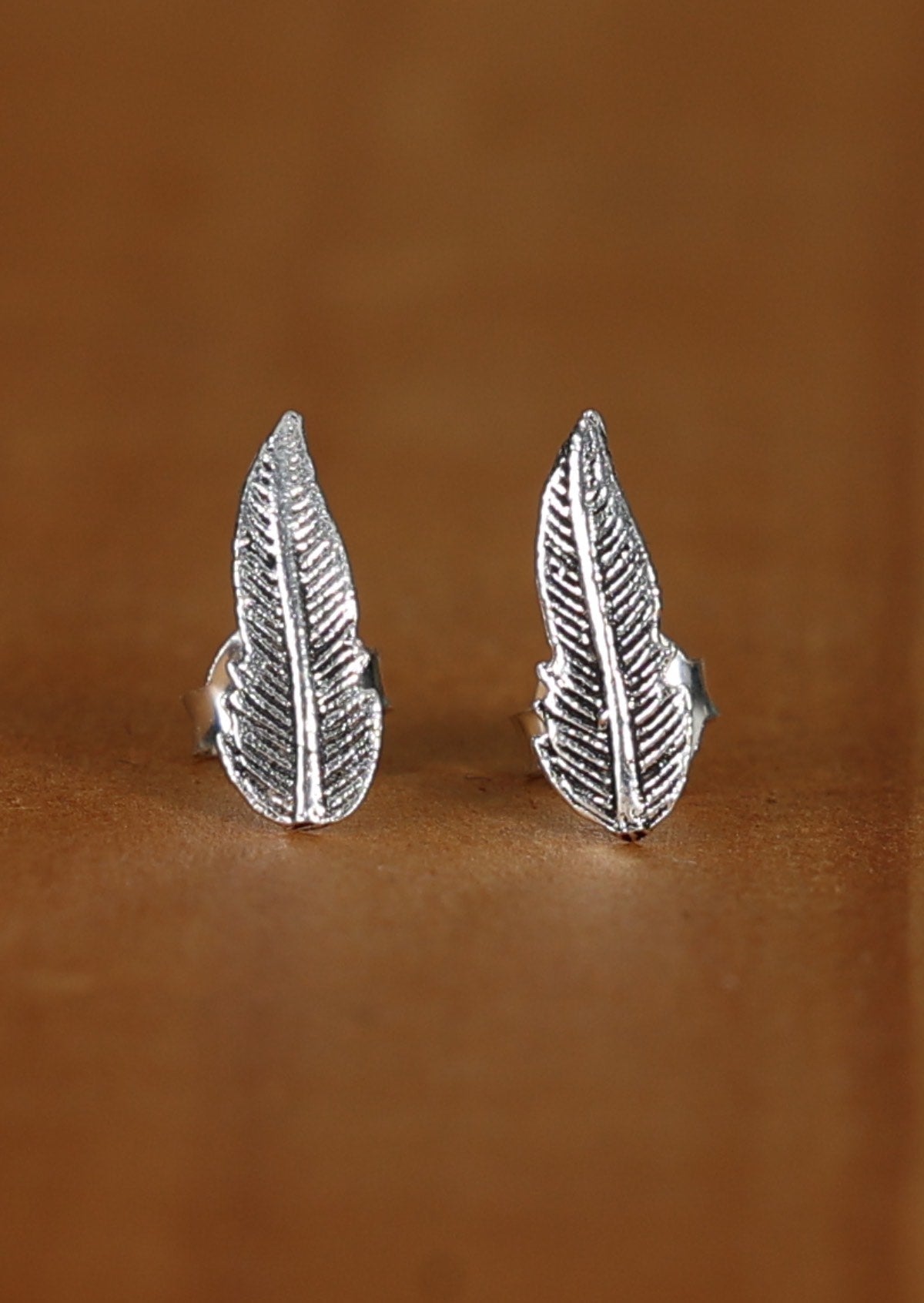 A pair of silver stud earrings with a feather design, displayed against a brown wooden background