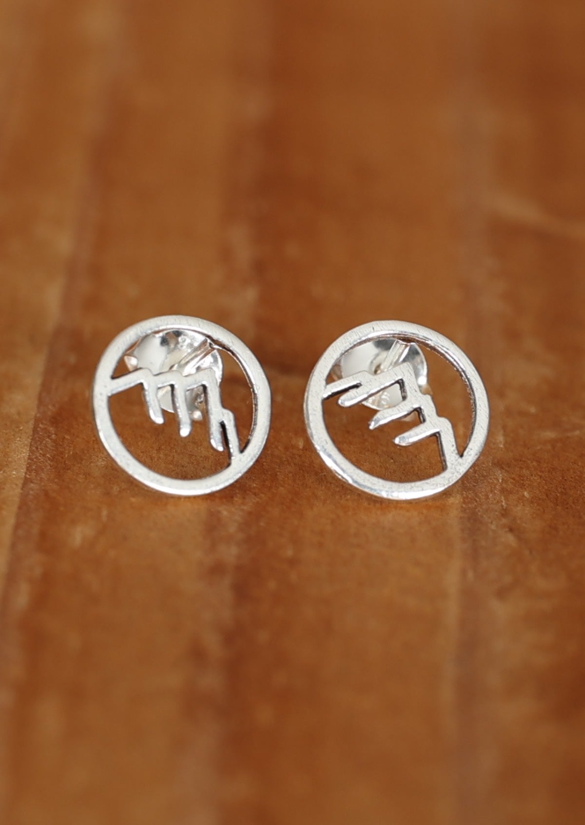 A pair of sterling silver Alp stud earrings with intricate mountain cutout details, displayed against a wooden background.