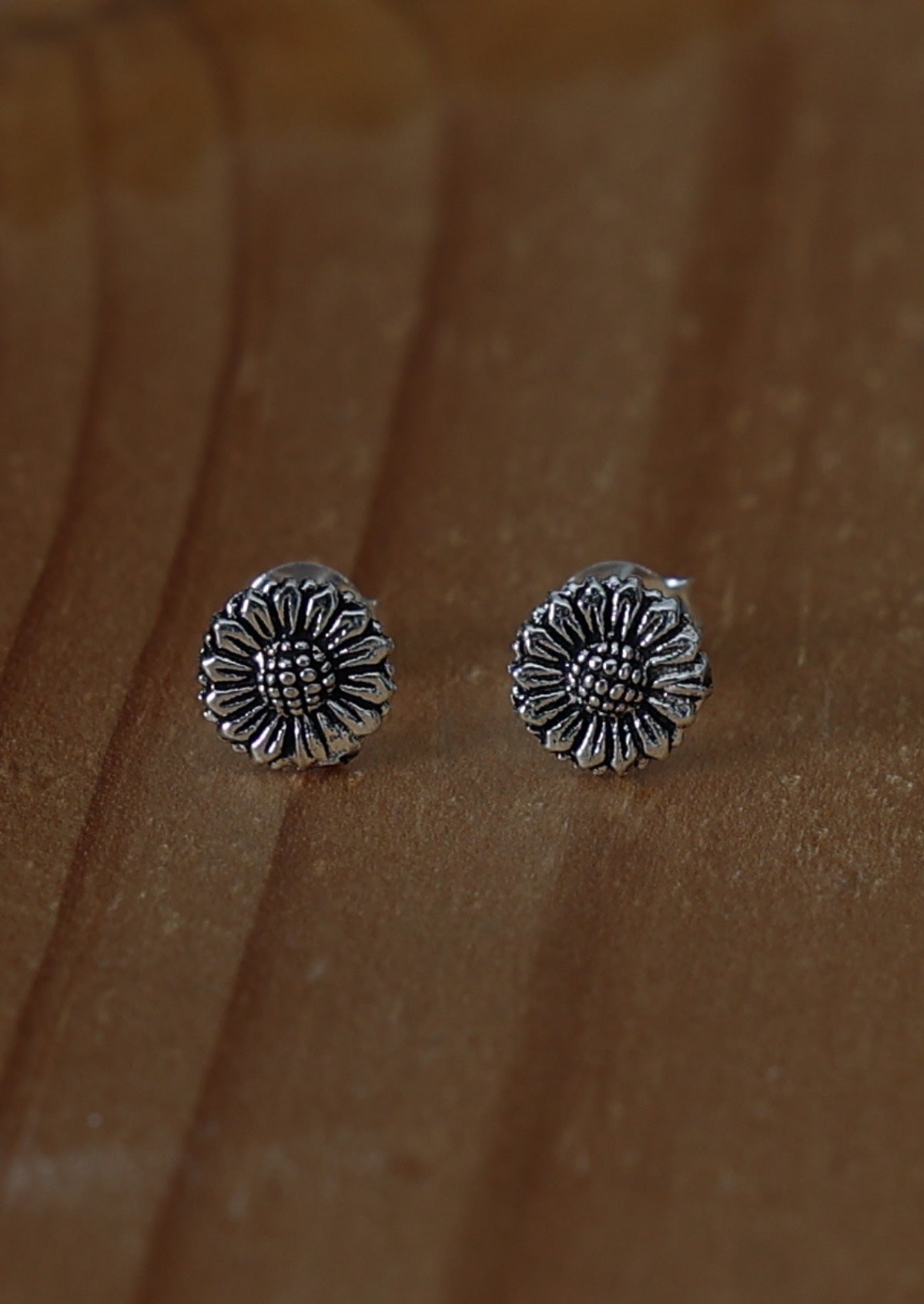 A pair of silver Radiant Sunflower stud earrings in the shape of a sunflower, with textured center and petals, displayed against a wooden background.