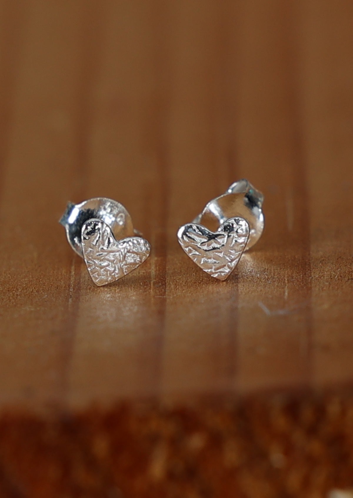 A pair of silver Dainty Heart stud earrings with a textured heart design, placed on a wooden surface.
