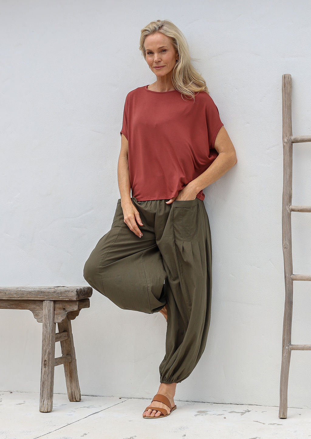 Woman standing in front of white wall wears loose fit cotton pants in olive green with deep pockets and elasticated ankles