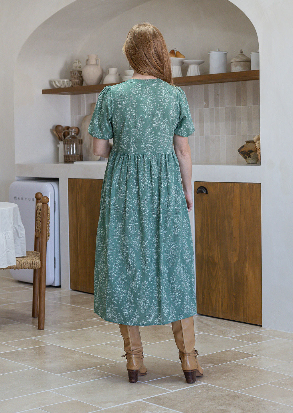 Woman standing backwards in a sage green based floral cotton button through dress with elbow length puffy sleeves and a V-neckline.