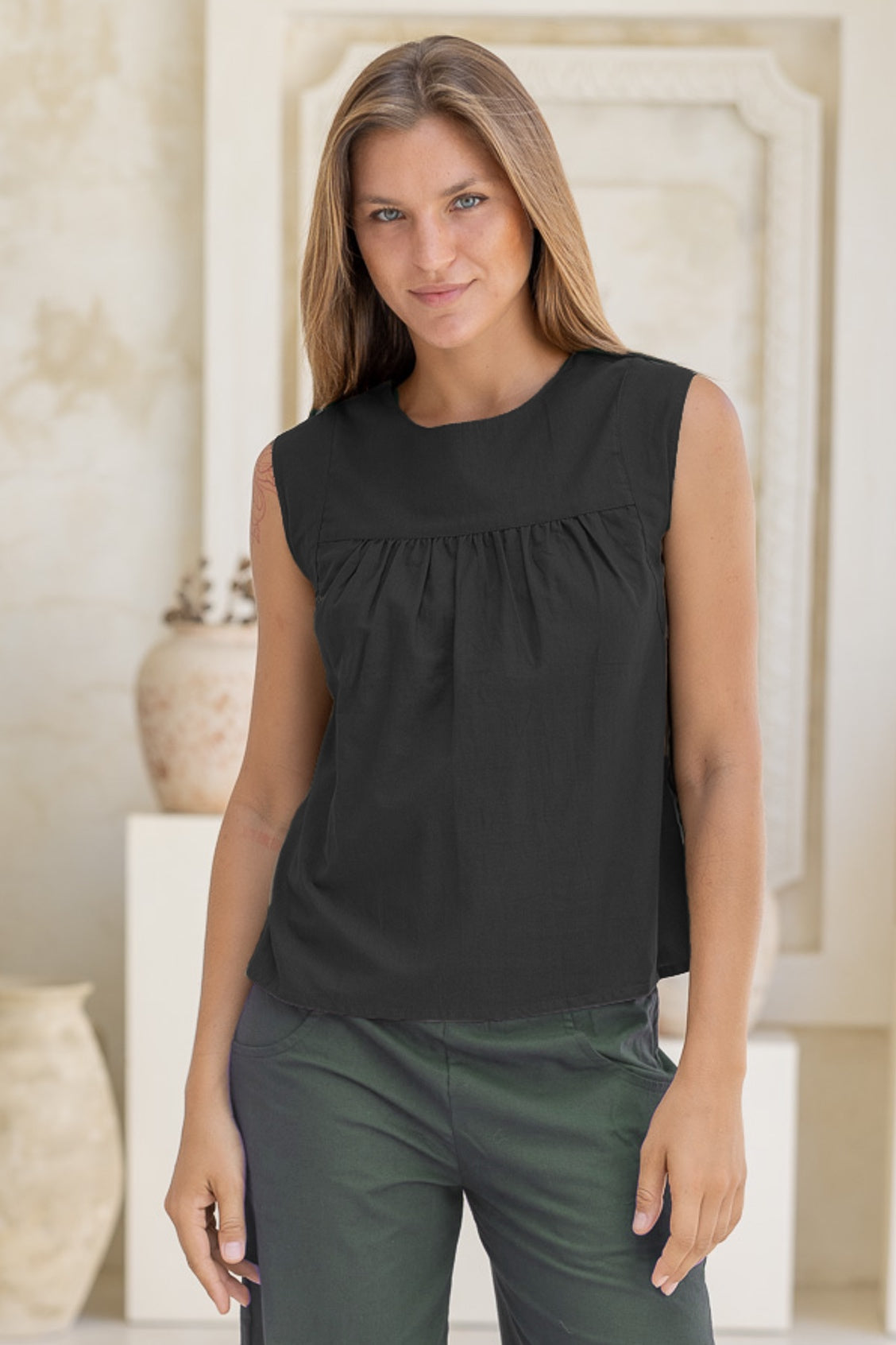 Woman wearing a black sleeveless cotton top with small gathers across the bust and green pants