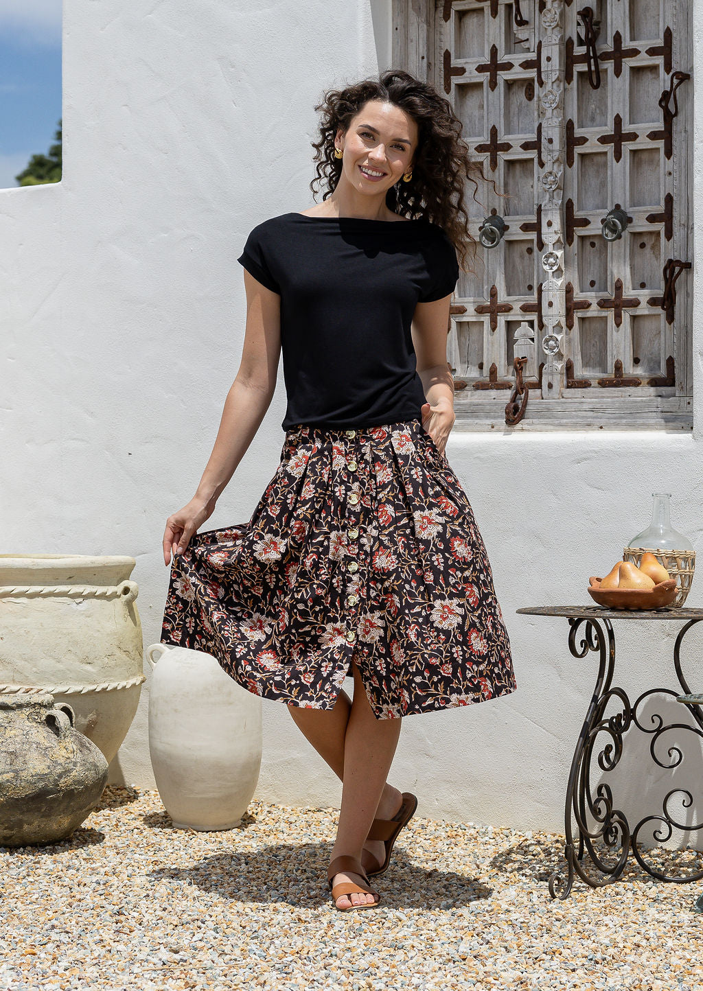 Woman stands wearing a basic black top and black based floral print, button through cotton skirt with box pleats and belt loops