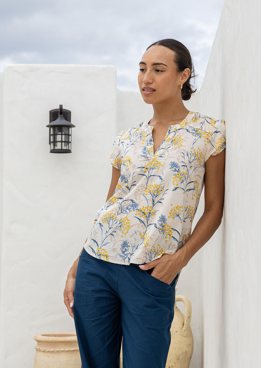 Woman wearing a neutral based floral blouse and blue pants standing against a white wall.
