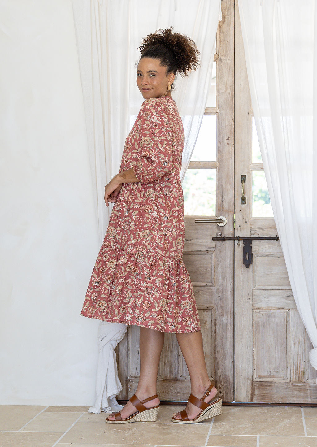 Model stands side on in cotton over the knee length relaxed fit dress with 3/4 sleeves, paired with tan sandals