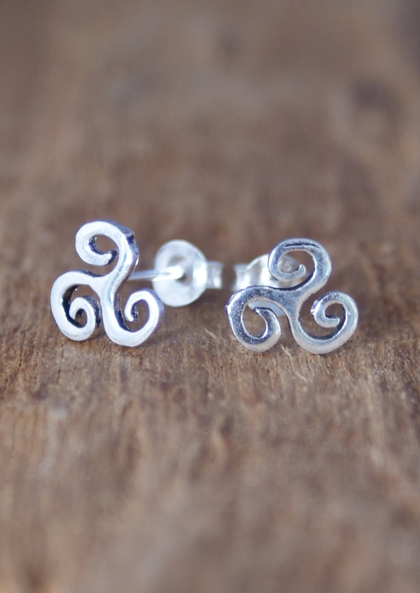 A pair of silver stud earrings with a triskele design, placed on a wooden surface.