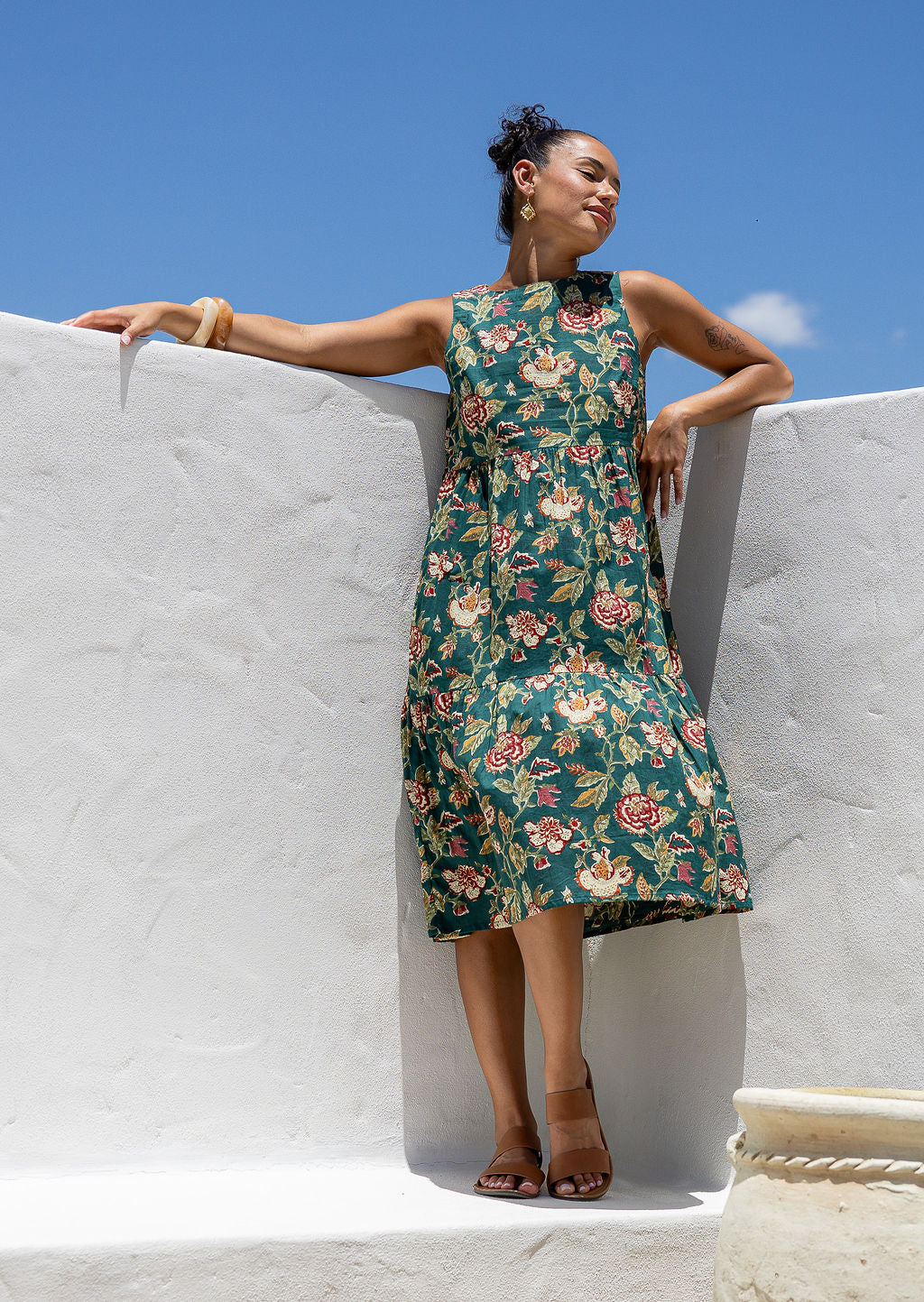 Woman in a sleeveless, floral green based 100% cotton dress standing in front of white wall.