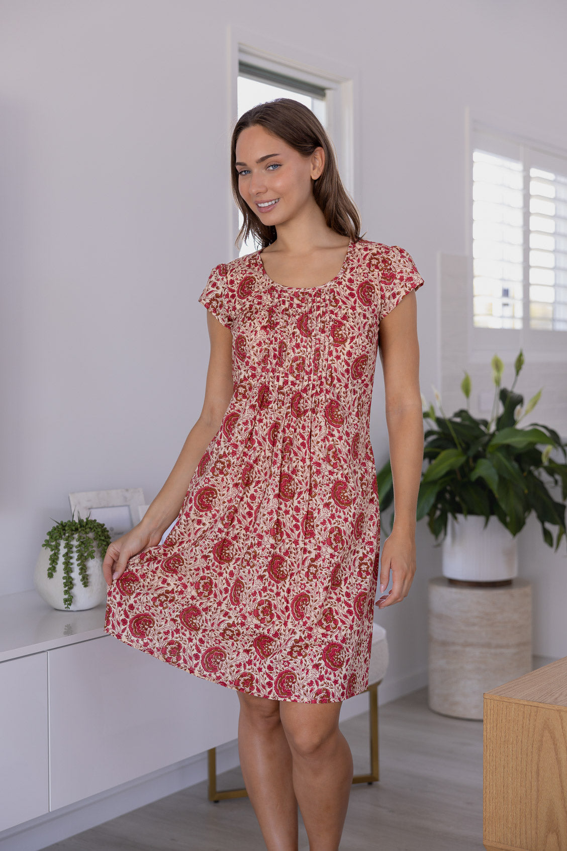 Woman wearing a 100% cotton gentle cream based floral dress in a modern room with white walls and plants.
