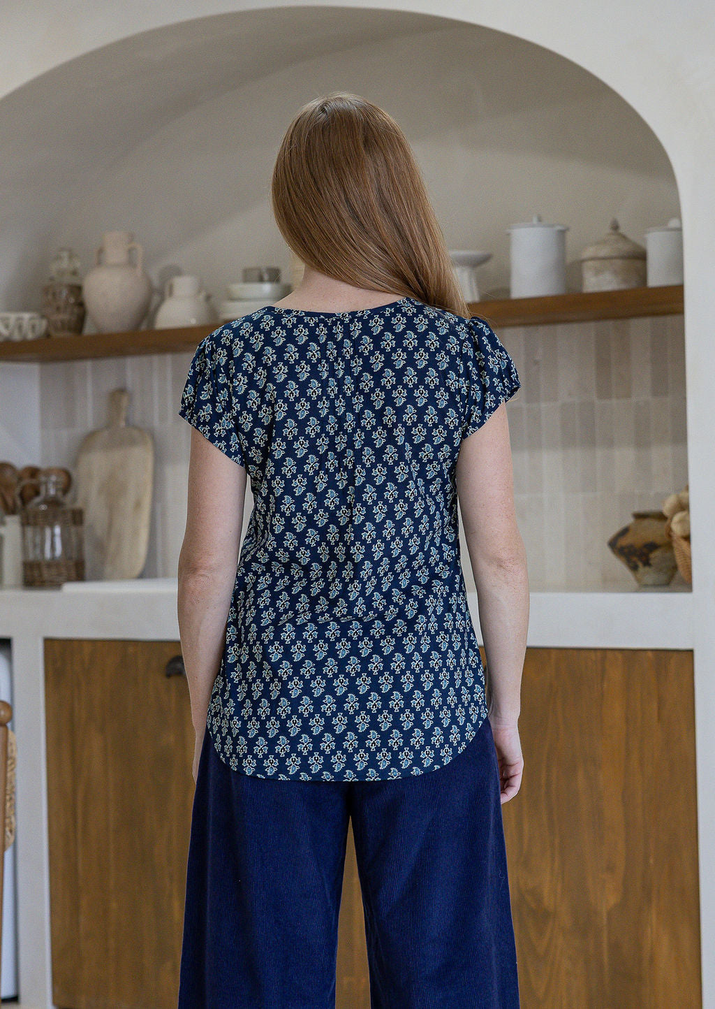 Woman stands backwards wearing 100% cotton top in navy blue based floral print with navy plain pants.