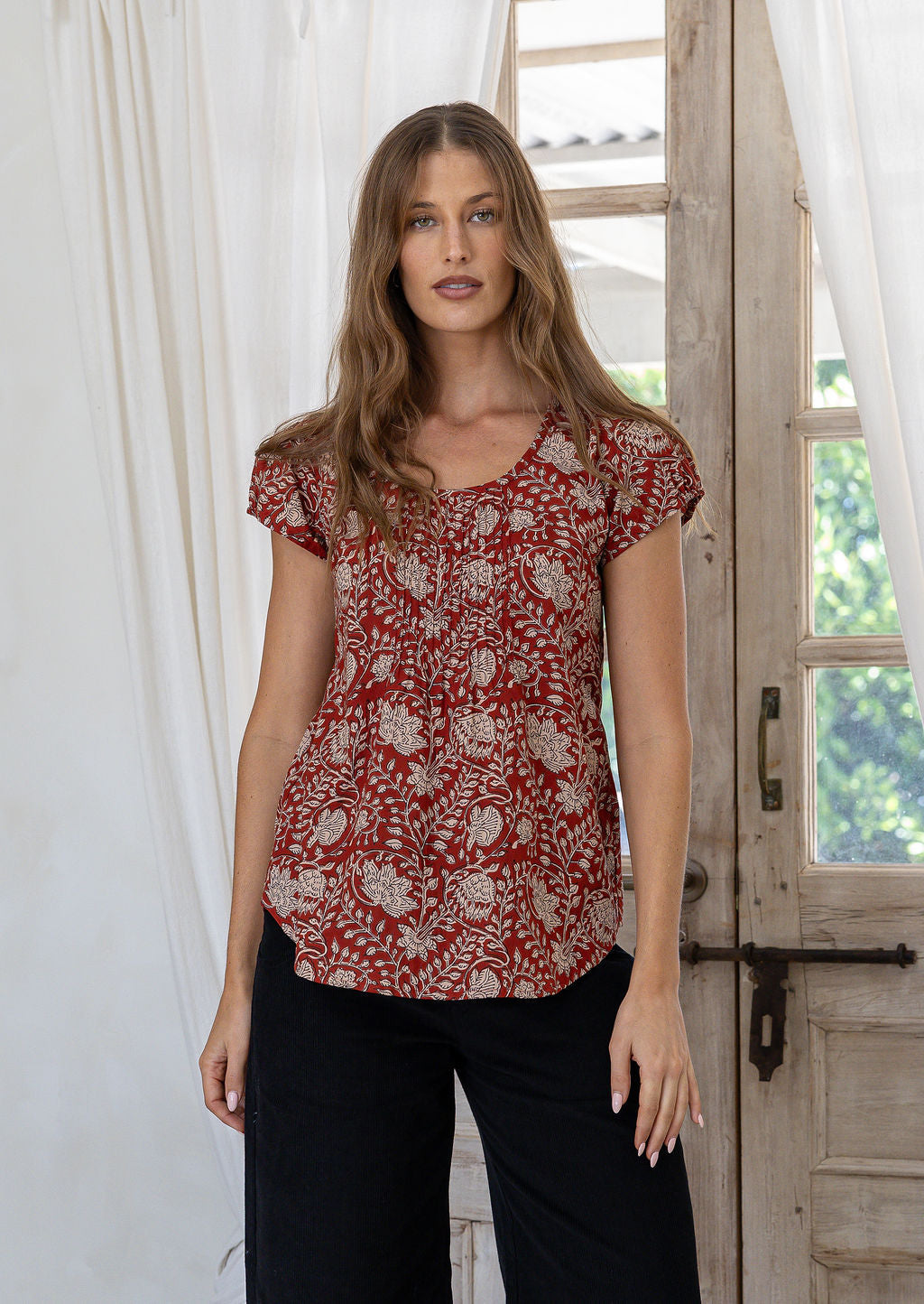 Woman wears red based floral print 100% cotton cap sleeve top with pin tuck detail across the bust and a scoop neckline