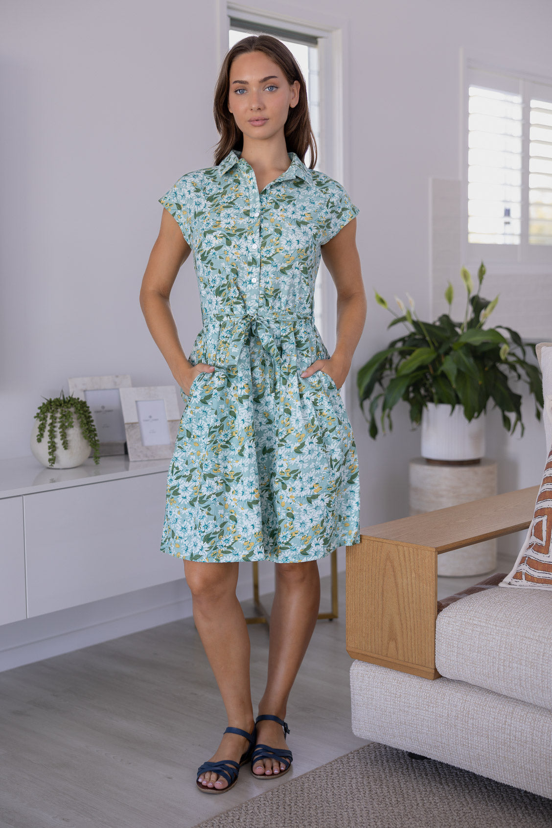 Model stands with both hand in pocket wearing a 100% cotton knee length shirt style retro floral dress in sage green based floral print.
