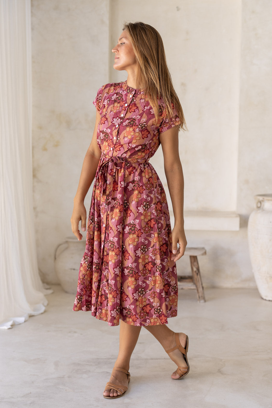 Woman stands slightly sideways wearing a cotton short sleeved dress with detachable waist tie and button through front in a warm pinkish toned floral print.