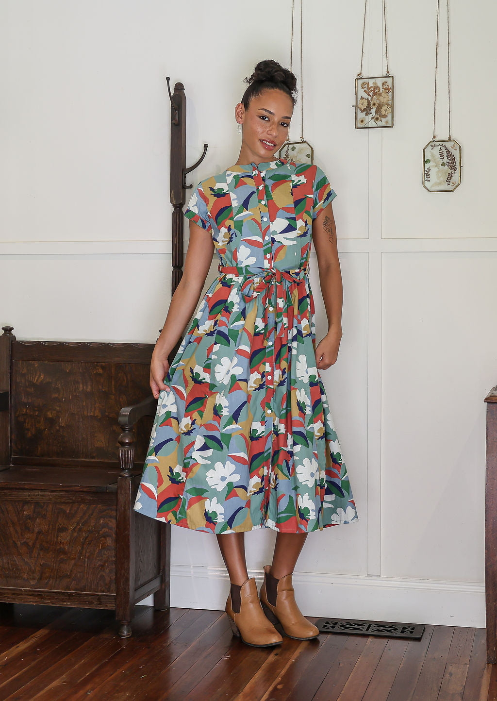 Woman wears retro style cotton button through midi length dress with high round neckline, short sleeves and a removable waist tie.