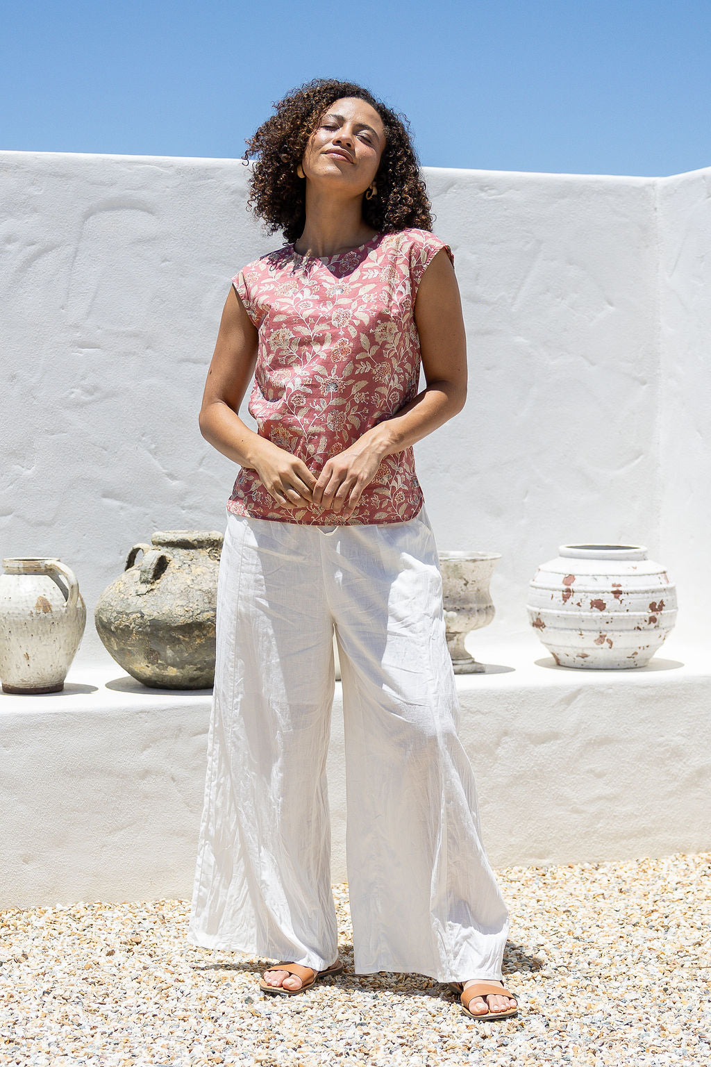 Woman wearing a warm pink based floral cotton top and white pants standing in front of a white wall