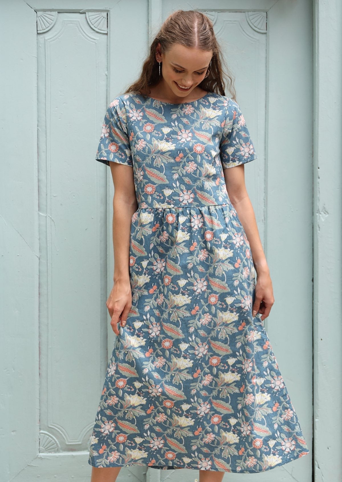 Woman stands in a short sleeve cotton calf length dress in a floral print with a mid blue base