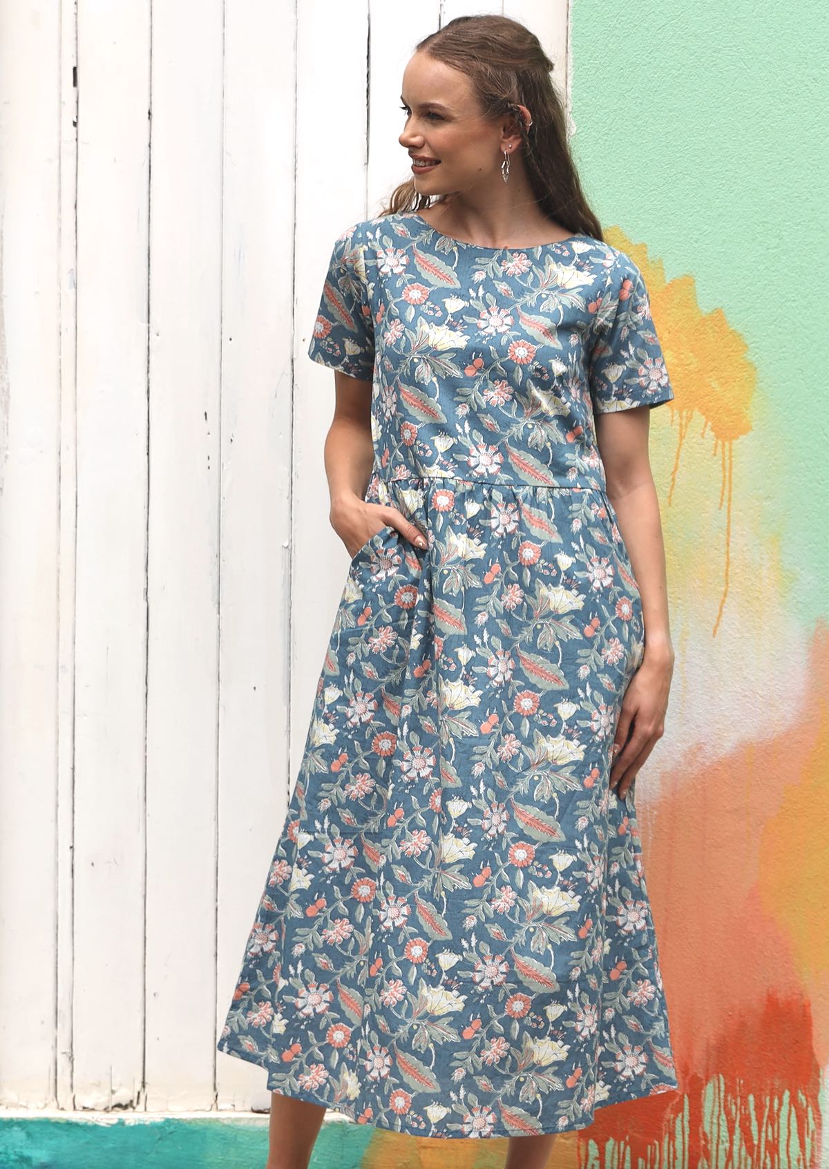 Woman wears mid blue based floral print cotton midi length dress with short sleeves and a relaxed waistline, with hand in pocket