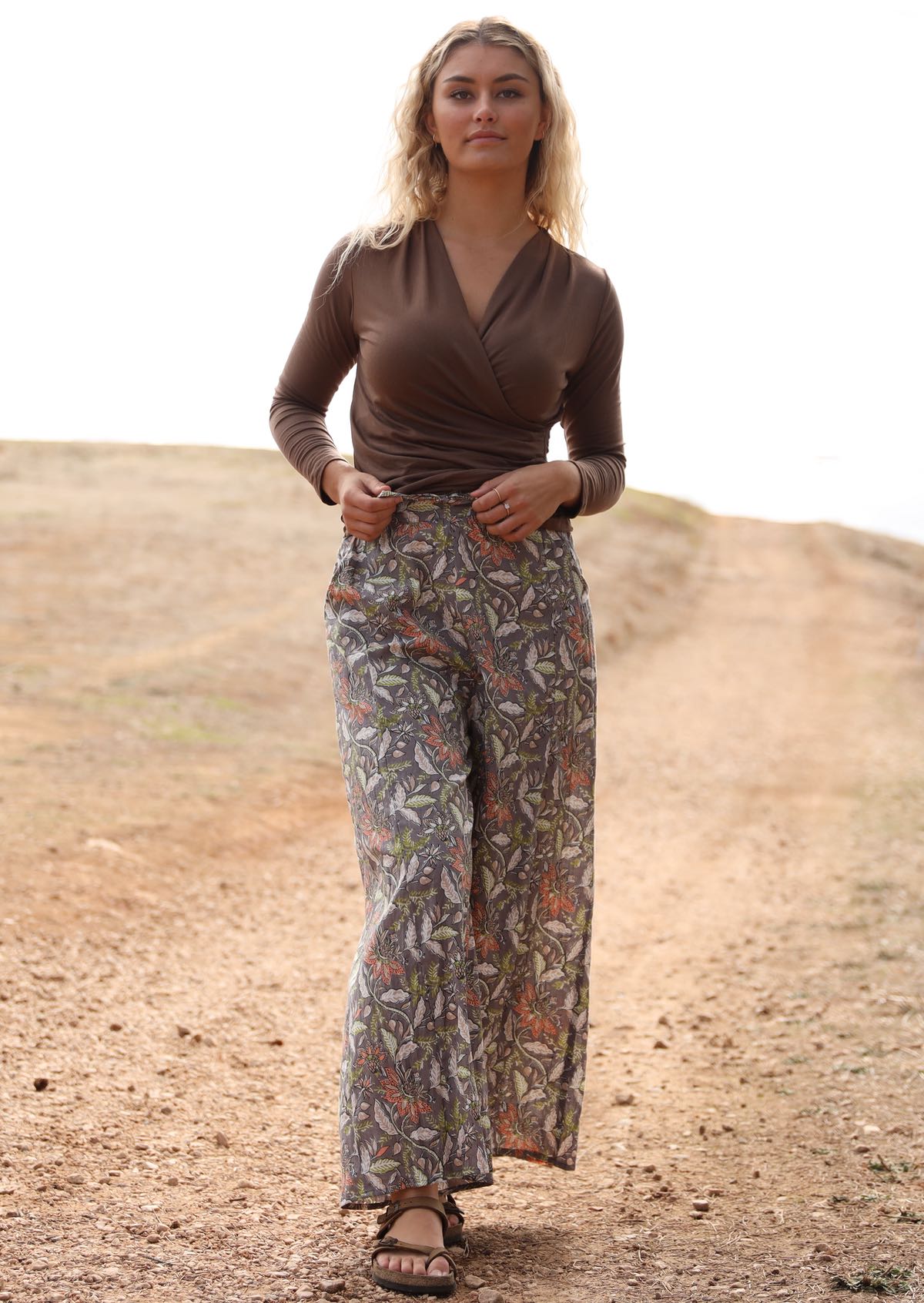 Woman wears lightweight cotton wide leg pants with taupe based floral print, paired with a brown false wrap top