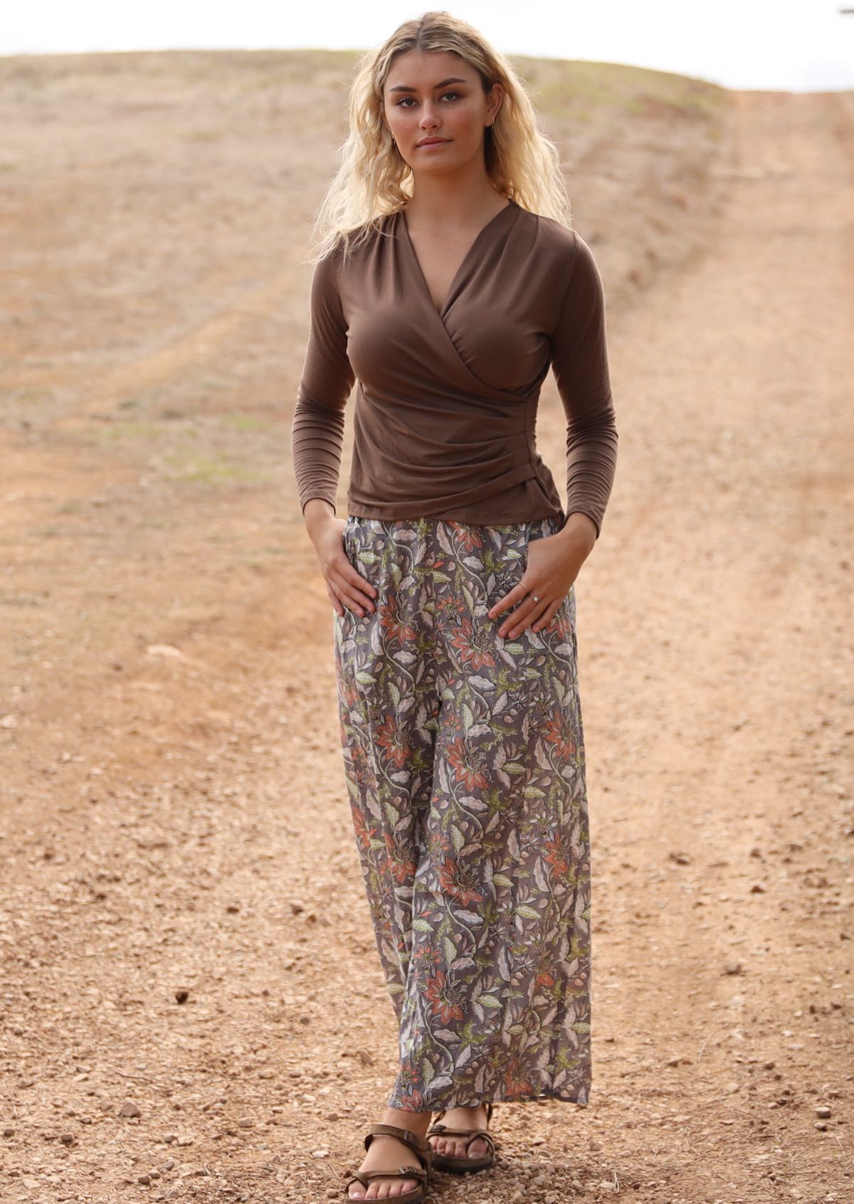 Woman walking down dirt road in cotton wide leg pants with taupe based floral print, paired with a brown top