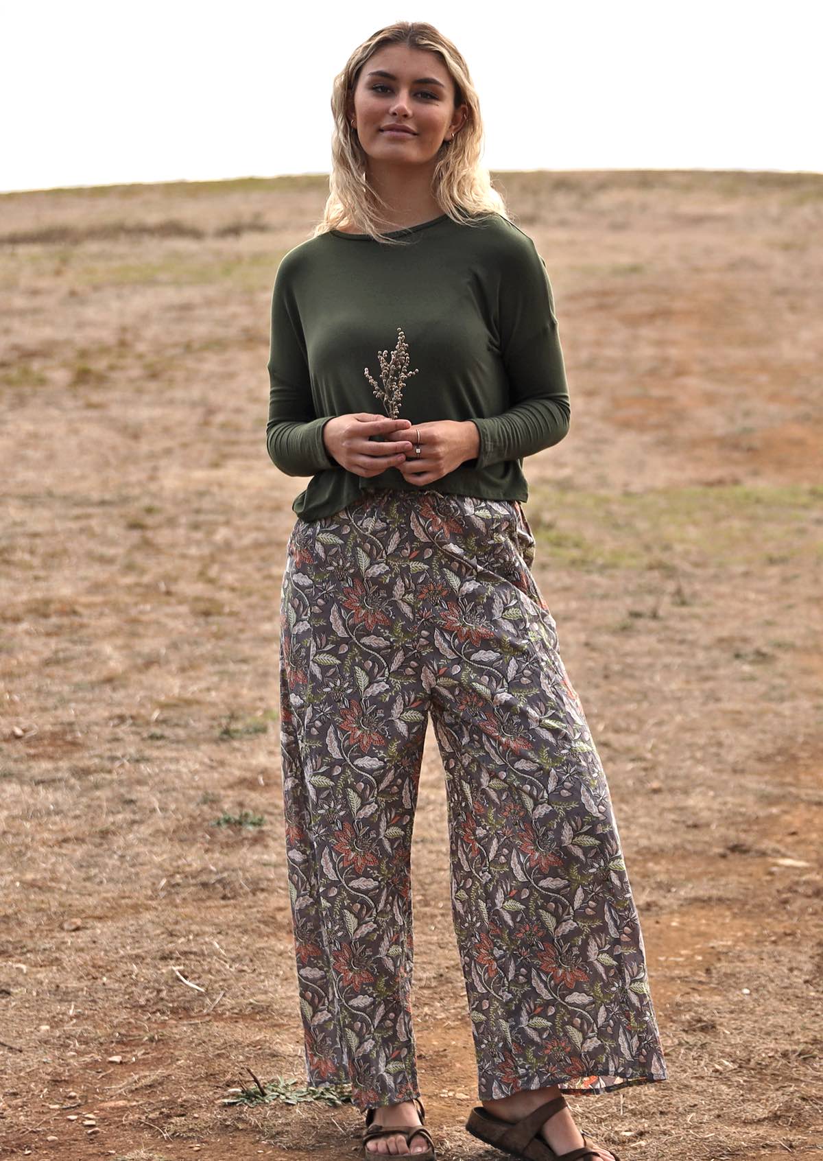 Woman wears cotton wide leg pants with taupe based floral print, paired with an olive green top, holding some small flowers