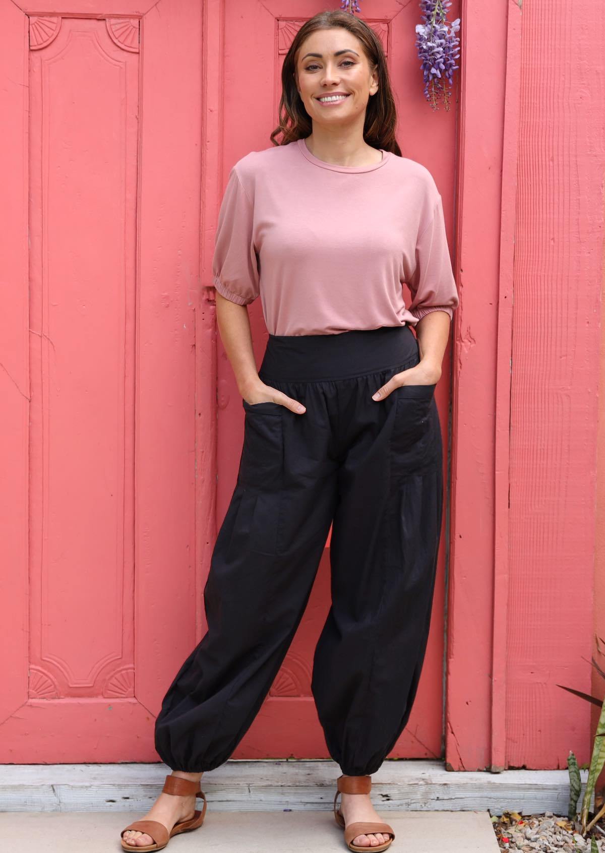 Woman wears lightweight black cotton harem pants paired with a dusty pink top and sandals, with hands in pockets