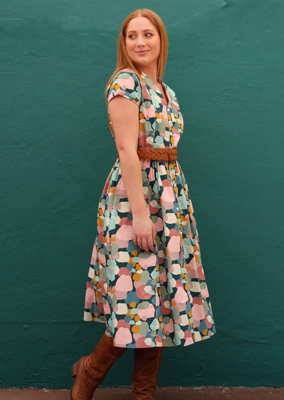 Model standing in front of dark teal background wears colourful retro style dress with boots and stylish belt at the waist.