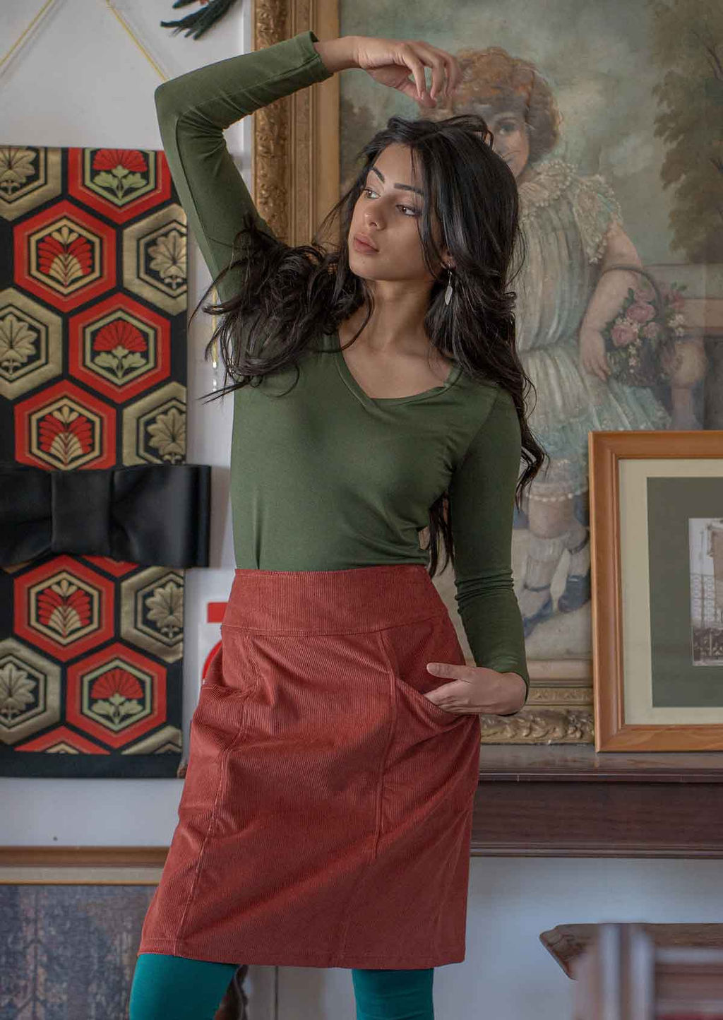 Woman raises hand over head as she poses in olive top with corduroy knee length skirt with one hand in pocket