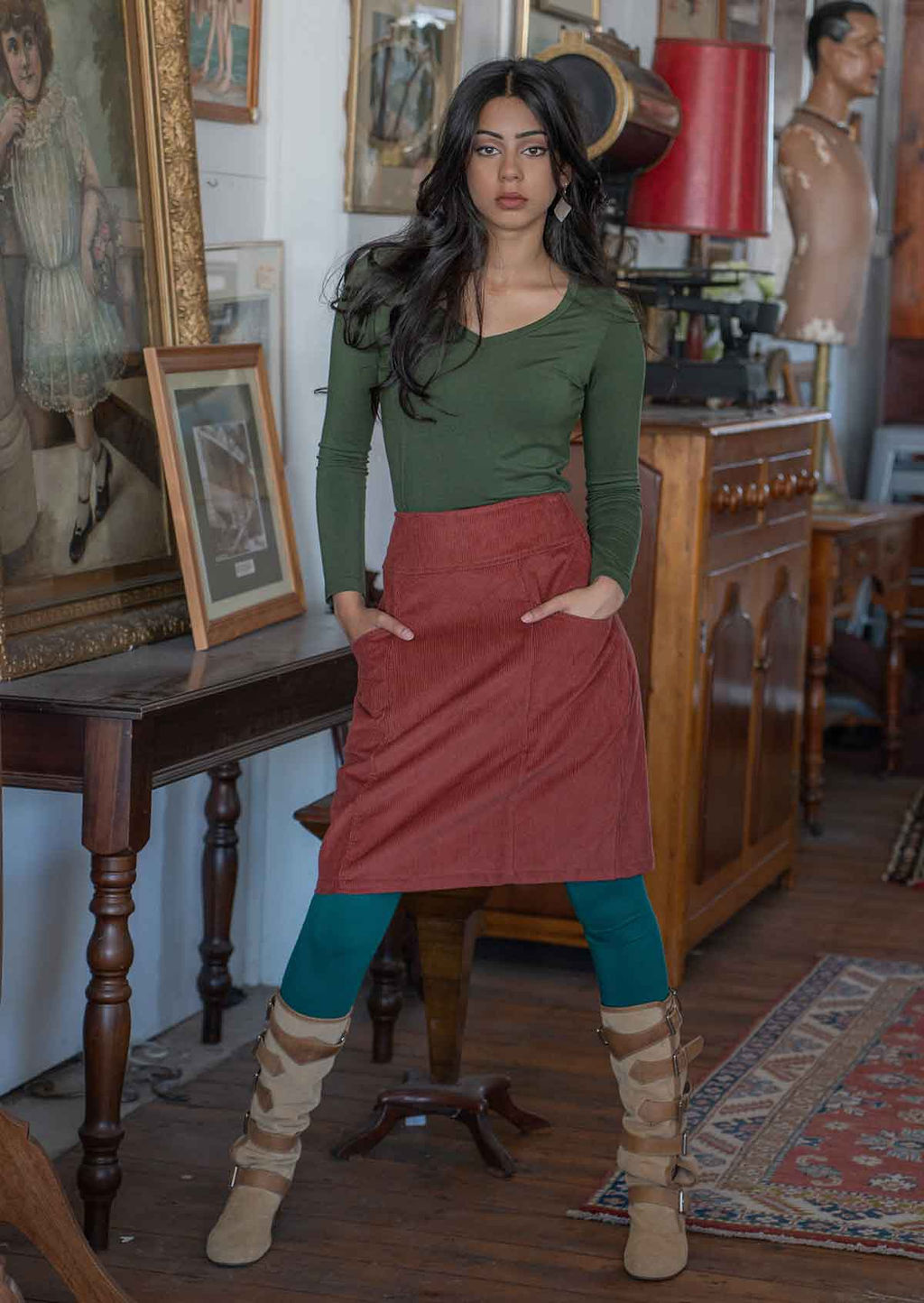 Model stands in A-line burnt henna corduroy skirt with her hands in pockets, paired with an olive long sleeve top and teal leggings