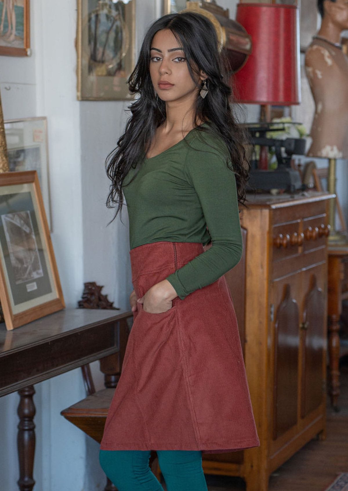 Woman stands sideways wearing a cotton corduroy A-line skirt with side zip with her hands in the pockets, and a long sleeve olive top