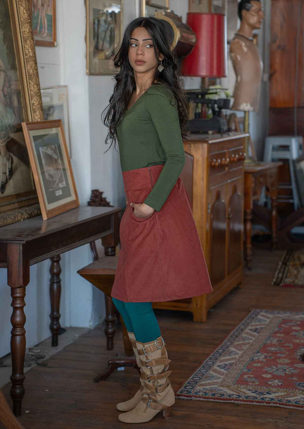 Woman poses side to the camera in an A-line knee length burnt henna skirt, paired with an olive top and teal leggings