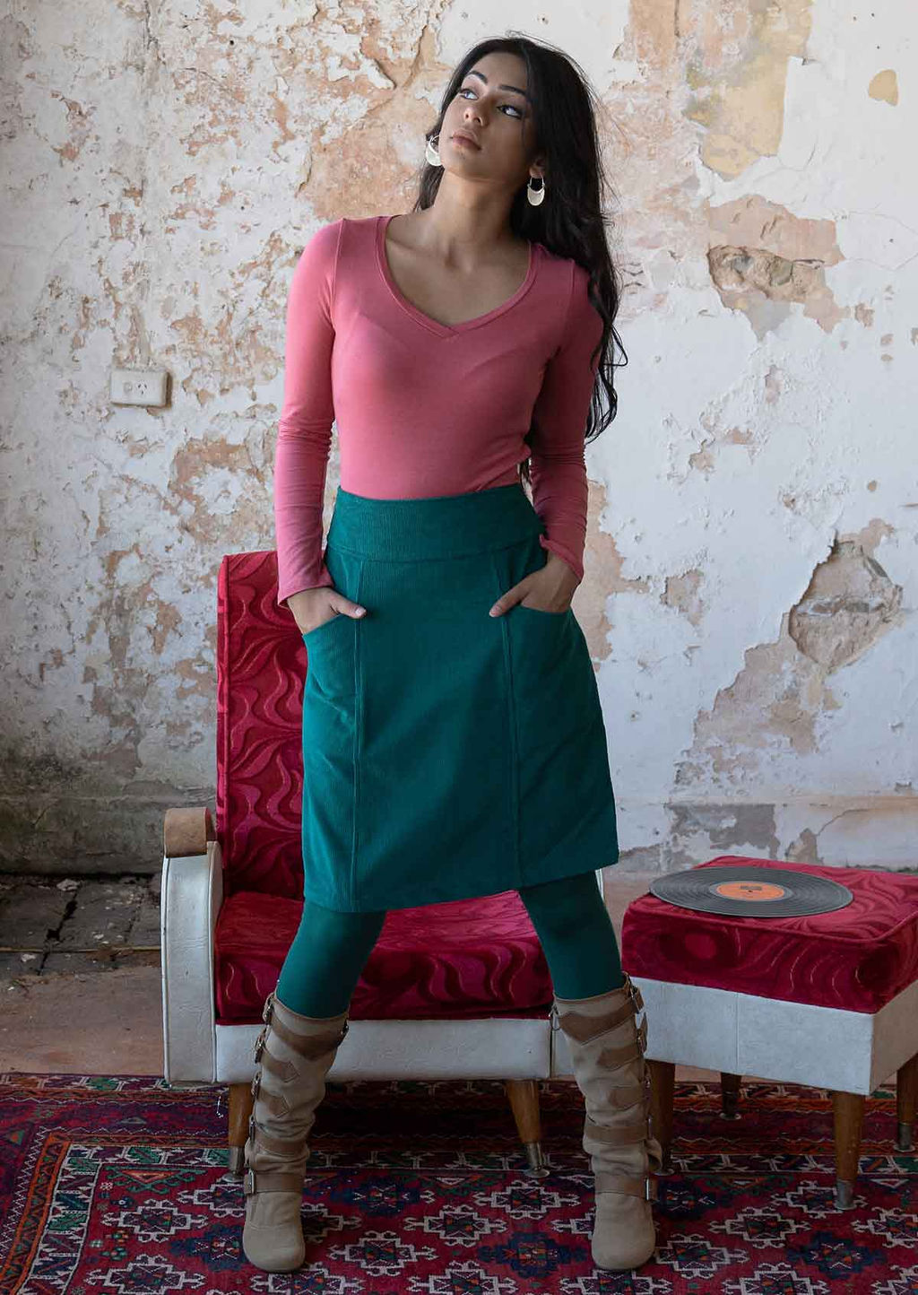 Woman wears cotton corduroy A-line knee length skirt in teal with hands in pockets, paired with a pink long sleeve top, leggings and boots