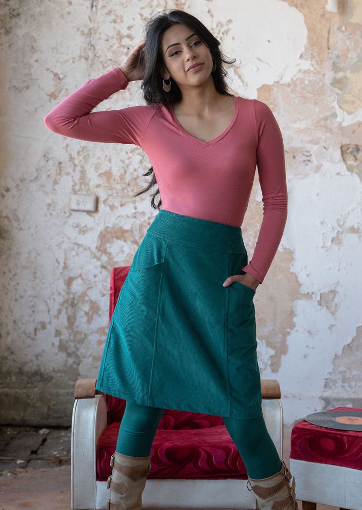 Woman wears corduroy knee length skirt in teal with hand in pocket, paired with a pink long sleeve top