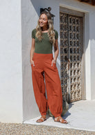 Woman wearing a olive green basic top and rust orange pull on cotton pants with wide leg fit and cuffed ankles standing against a white wall with a wooden gate.